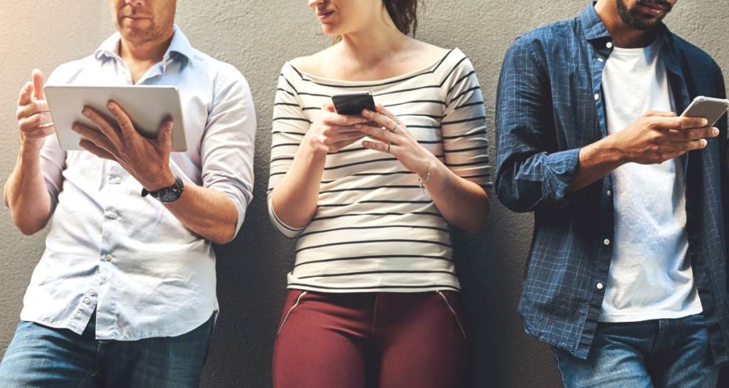 Application Performance Monitoring (APM) na prática: monitoramento contínuo para alta disponibilidade três pessoas encostadas em uma parede, cada uma usando um dispositivo digital, um tablet e dois smartphones.