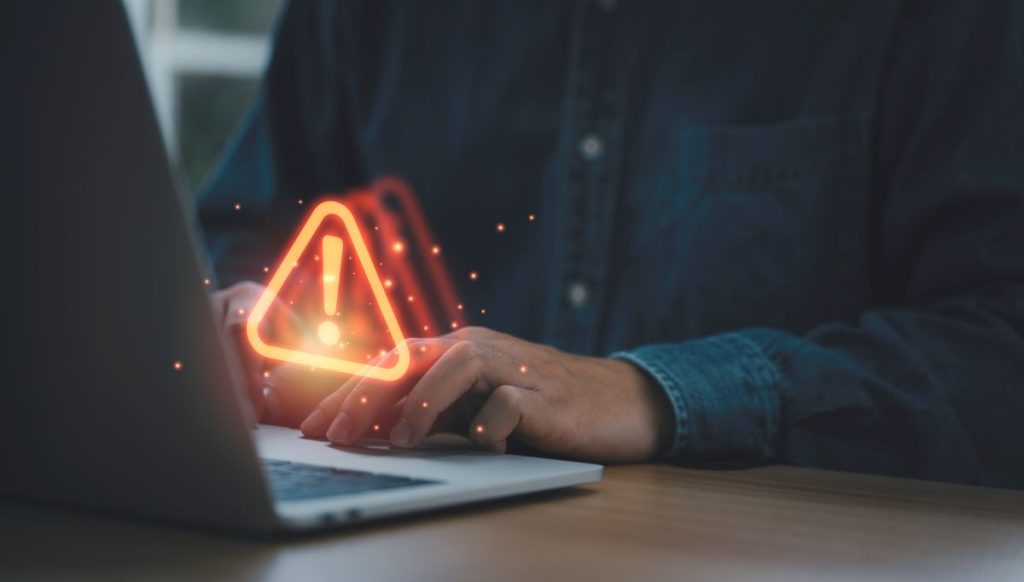 : Uma imagem mostra uma pessoa usando um laptop, enquanto um ícone luminoso de alerta com um ponto de exclamação aparece no teclado. 